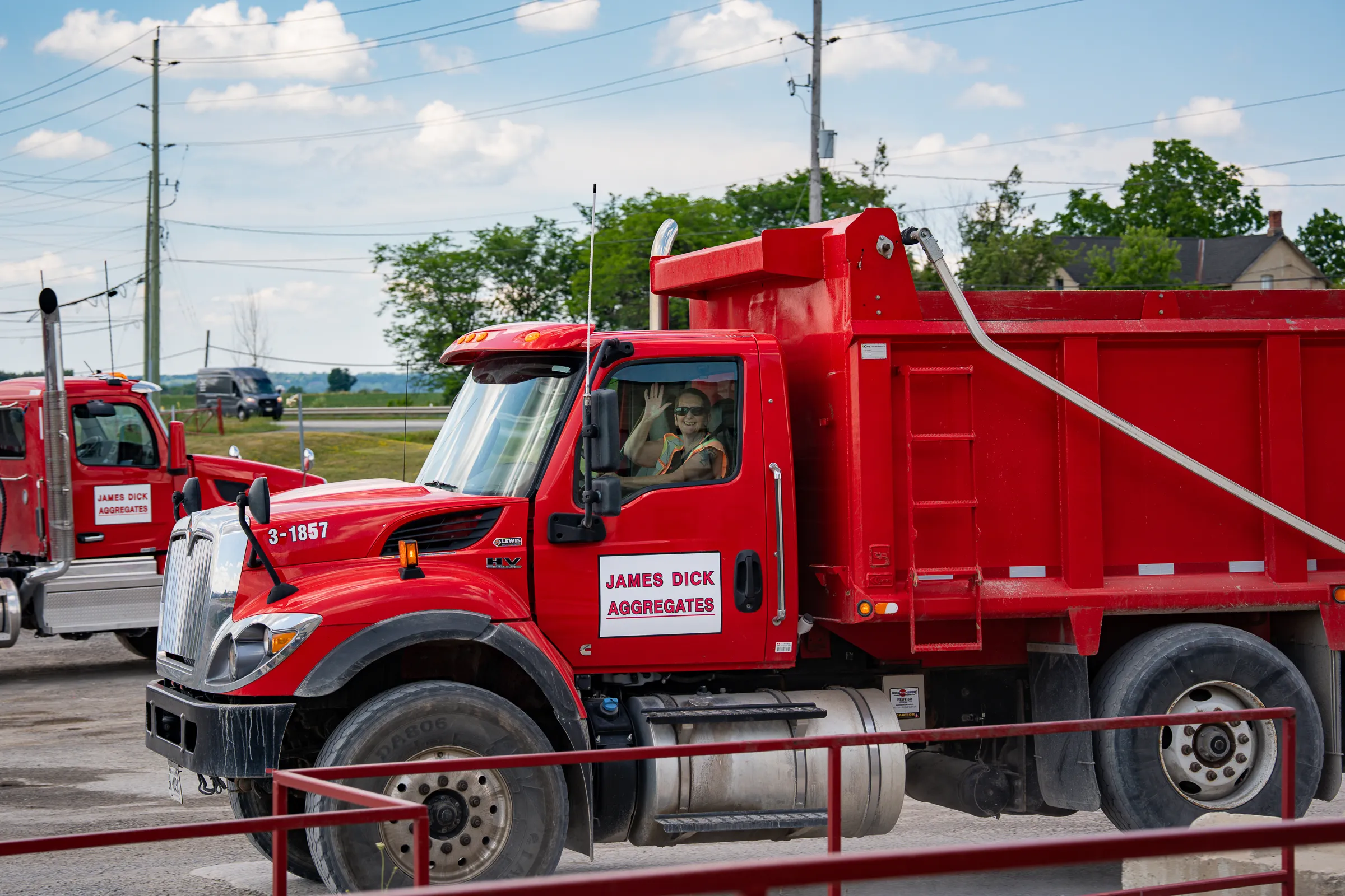 James Dick Aggregates driver in high-visibility vest waves from the cab of a red JDA dump truck at the yard