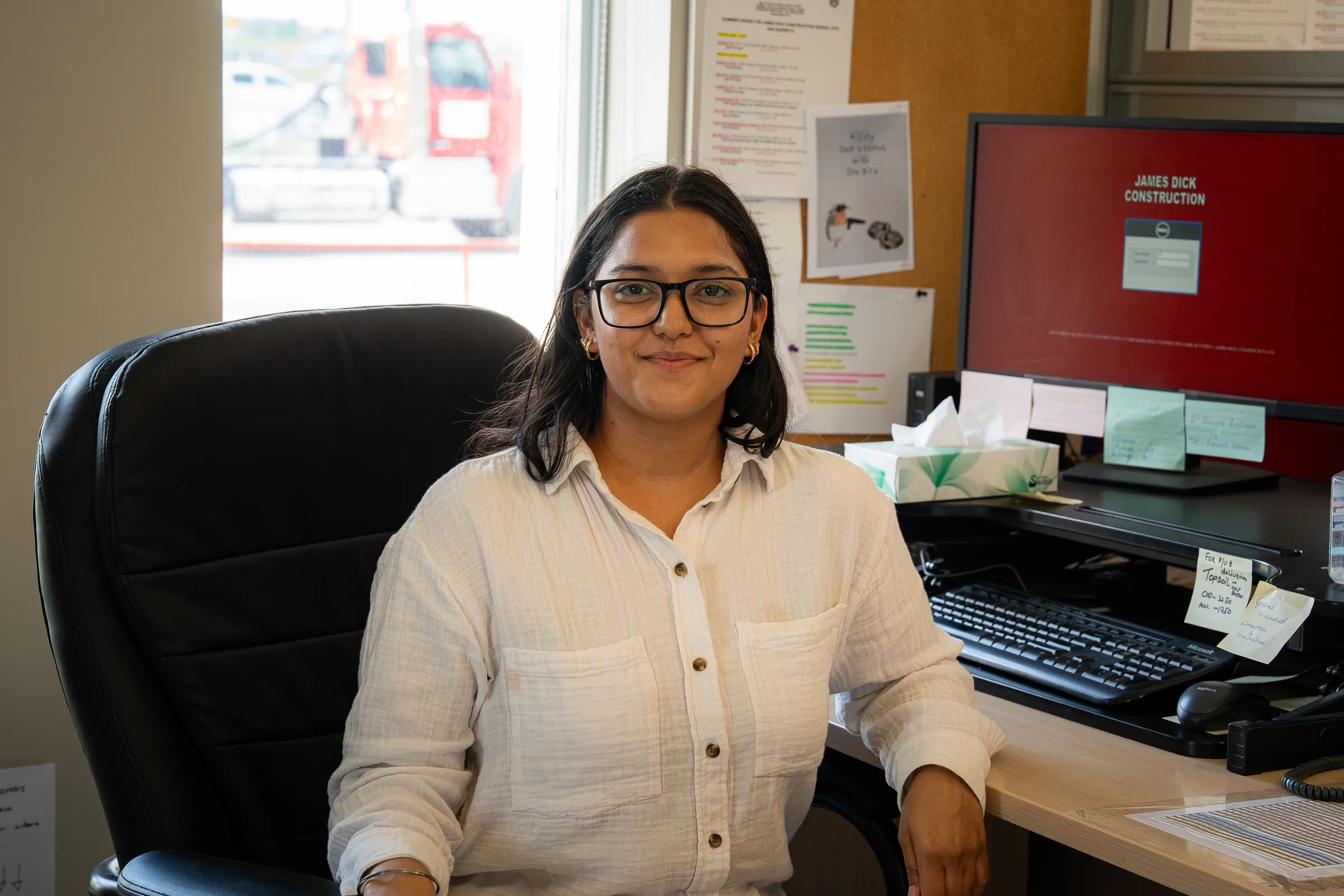 JDA office team member at her desk with a computer and bulletin board, a red JDA truck visible through the window behind her